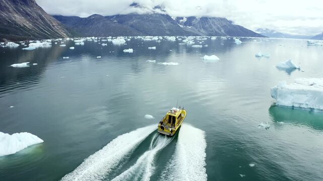 Aerial view of a yellow boat sailing across the water, contrasting with white wakes and floating icebergs, near mountains, Ilulissat, Avannaata, Greenland.