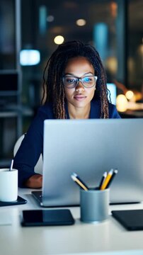 An African-American woman with glasses and dreadlocks is working at her desk. She is looking at her laptop screen and typing late at night in the office