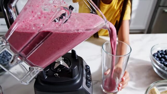 Girl adding fruits to blender and pouring smoothie into glass. Bright kitchen with modern design and fresh produce on countertop. Concept of healthy living, cooking at home, family meals
