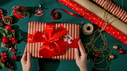 Top view of female hands holding a beautifully wrapped Christmas gift in striped paper with a shiny red satin bow. Festive decorations surround the gift, creating a warm and joyful holiday atmosphere.