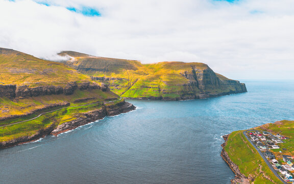 Aerial view of dramatic cliffs meet the sea, a village nestled at the water's edge, lush green hills contrasting with the dark ocean, Tj&Atilde;&cedil;rnuv&Atilde;&shy;k, Streymoy, Faroe Islands.