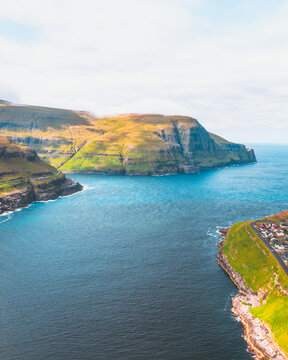 Aerial view of the rugged coastline meeting the deep blue sea, where the small village nestles at the foot of the towering cliffs, Tj&Atilde;&cedil;rnuv&Atilde;&shy;k, Streymoy, Faroe Islands.