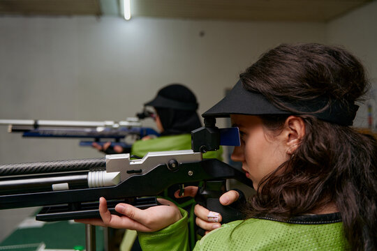 Focused female athletes aiming with air rifles, precision shooting competition, indoor range, concentration and skill, competitive sport, professional training, target practice