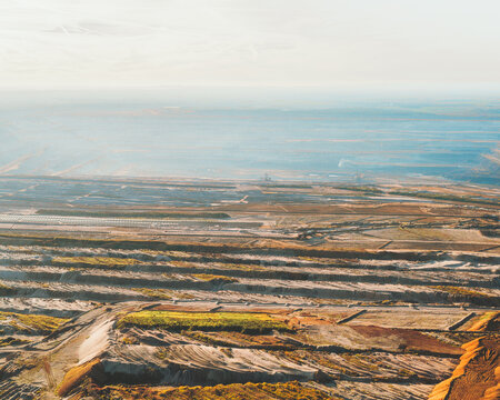 Aerial view of the vast, scarred landscape of Tagebau Hambach, a testament to human intervention, with layered earth tones contrasting against the pale sky, Elsdorf, Nordrhein-Westfalen, Germany.