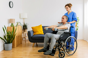 Old man in wheelchair training with physiotherapist using dumbbells at home. Female therapist assisting senior man with exercises in nursing home service