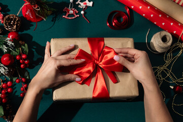 Female hands adjusting a red satin ribbon bow on a kraft paper wrapped Christmas present surrounded by festive decorations, and wrapping materials on a green background.