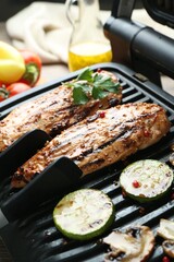 Electric grill with tasty chicken fillet, mushrooms, zucchini and tongs on table, closeup