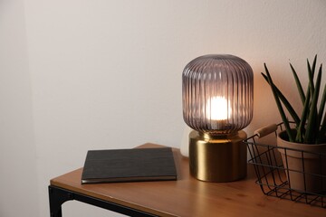 Stylish lamp, houseplant and book on wooden table near white wall indoors, space for text