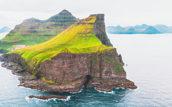 Aerial view of dramatic cliffs rise, adorned with vibrant green vegetation against the dark rock, contrasting with the serene blue sea, Kallur Lighthouse, Nor&Atilde;&deg;oya, Faroe Islands.