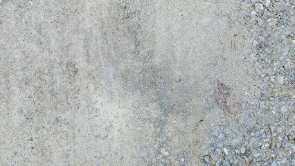 Rough concrete surface with scattered pebbles and a single dry leaf, featuring a mix of gray, beige, and earthy tones that highlight the natural texture and uneven details.