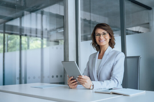 European businesswoman CEO holding digital tablet using fintech tab application sitting at workplace in office. Latin Hispanic mature adult professional business woman looking at camera. Copy space
