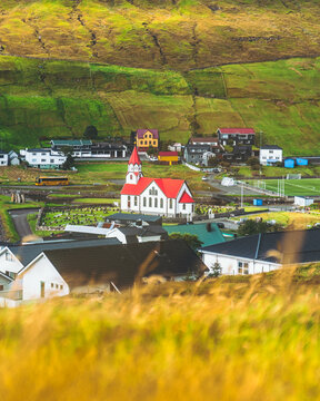 View of a quaint village nestled amongst vibrant green hills, punctuated by a red-roofed church, creating a serene yet striking vista, Sandav&aacute;gur, V&aacute;gar, Faroe Islands.