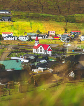 View of a charming village nestled in green hills with a red-roofed church as a focal point, houses scattered across the landscape, Sandav&aacute;gur, V&aacute;gar, Faroe Islands.