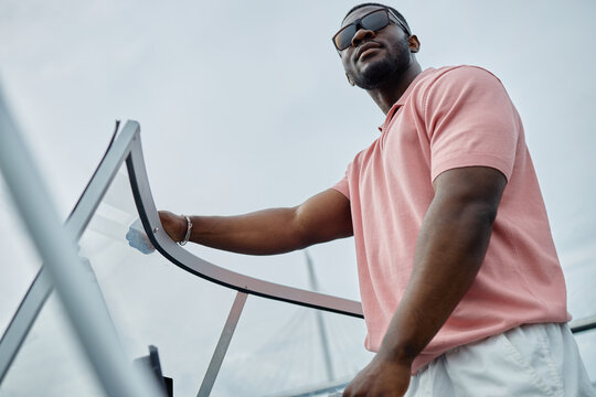 Black young adult man steering boat outdoors, wearing sunglasses, standing at helm with confident posture, looking away from camera, enjoying leisure activity on water
