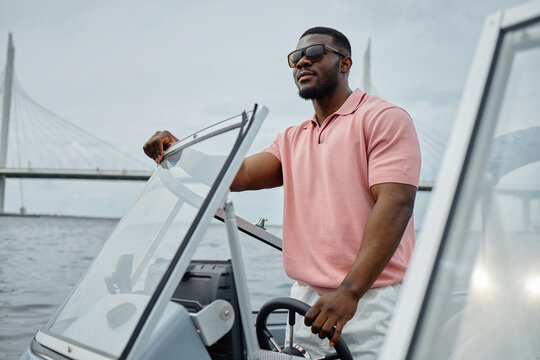 Black young adult man steering motorboat on water, wearing sunglasses, confidently navigating near modern bridge, standing at helm with one hand on wheel, looking ahead