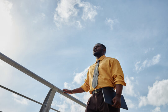 Young adult Black man standing outdoors holding laptop, looking into distance with confident expression, blue sky and clouds in background, business professional setting