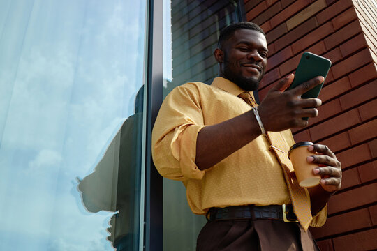 Black young adult man standing outdoors holding smartphone in one hand and disposable coffee cup in other hand, smiling while looking at mobile device near glass building