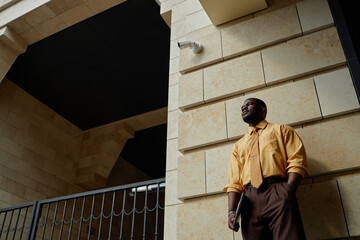 Black young adult man standing outdoors near modern stone building holding laptop, looking away with thoughtful expression, business professional in urban environment