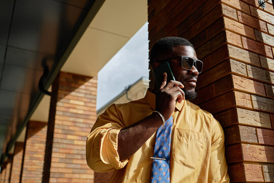 Young adult Black man standing outdoors leaning against brick wall talking on smartphone wearing sunglasses and tie looking away from camera with confident expression