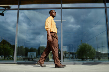 Young adult Black man walking confidently outdoors in front of modern glass building, wearing sunglasses, looking ahead with relaxed posture, urban environment reflected in windows