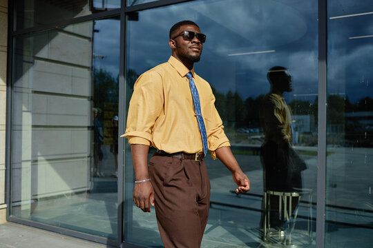 Young adult Black man walking outdoors near glass building, wearing sunglasses and smiling, confident expression, modern urban setting reflected in background window