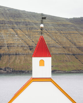 View of a quaint white church with a striking red roof piercing the skyline against a backdrop of dramatic cliffs, Mikladalur, Northern Isles, Faroe Islands.