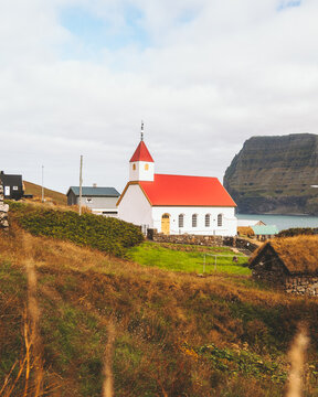 View of a striking white church with a vibrant red roof stands proudly amidst the windswept, golden hills, overlooking the serene, deep blue sea, Mikladalur, Northern Isles, Faroe Islands.