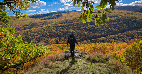 A lone hiker stands on the edge of the Vidróczky Lookout in the Bükk Mountains, Hungary, surrounded by colorful autumn forest and dramatic hills under a bright blue sky.