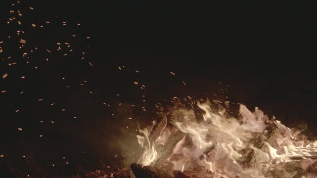 Slow-motion footage of a bonfire at night as a log is thrown onto the fire, sending sparks and embers upward while the camera pans toward the rising glow, filmed outdoors in rural England.