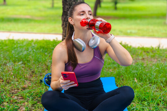 Sporty young woman sitting on mat in park after exercising, drinking water from red bottle while holding smartphone, wearing headphones around her neck and fitness outfit, relaxing under daylight.