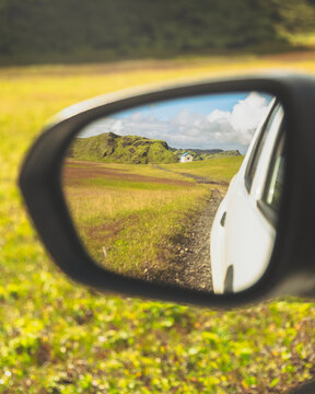 View of a car's side mirror reflecting a vibrant landscape of rolling green hills under a partly cloudy sky, a scenic vista, F208, Iceland.