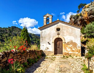 Fototapeta premium A charming, weathered stone chapel sits nestled amongst lush greenery and rugged cliffs under a bright blue sky. A cobblestone path leads to its doorway