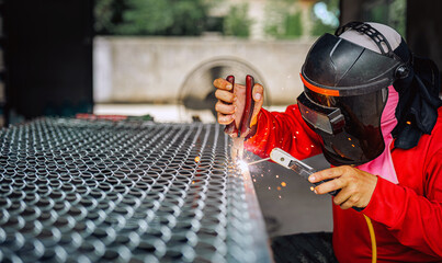 Welder wearing a red long-sleeved shirt and a protective mask is welding a steel grating, Industrial Welder With Torch