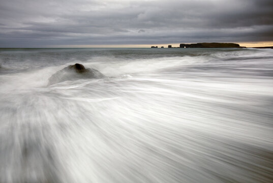 View of a dark rock battles the violent, foamy surf under a brooding sky, with distant cliffs silhouetted against the horizon, V&iacute;k, Myrdalshreppur, Iceland.