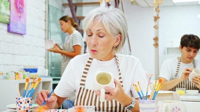 Interested elderly woman attending pottery class, enthusiastically painting on ceramic cup, creating unique custom design. Creative hobby concept