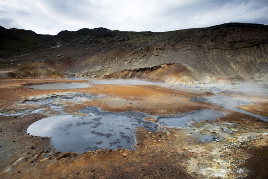 View of steaming geothermal pools reflecting the cloudy sky, surrounded by ochre and brown earth, creating a surreal landscape, Reykjanes, Grindav&iacute;kurbaer, Iceland.