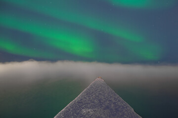 View of the snow-dusted triangular rooftop piercing the ethereal mist under the mesmerizing green glow of the Aurora Borealis, Grindavíkurbaer, Iceland.