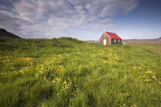 View of a quaint church with a vibrant red roof atop a grassy knoll dotted with yellow wildflowers under a cloudy sky, Grindav&iacute;kurbaer, Iceland.
