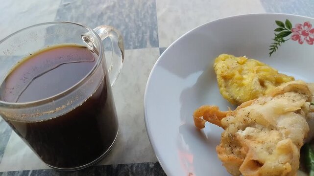 Close-up, high-angle shot of a glass mug of hot black coffee next to a plate of traditional Indonesian snacks (gorengan), specifically fried food like pisang goreng (banana fritters) and possibly frie