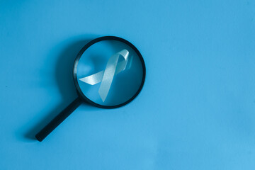 close up top view white ribbon over magnifier glass on blue background for symbol of cancer awareness and health care concept