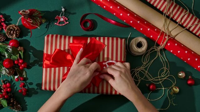 Top view of female hands holding a beautifully wrapped Christmas gift in kraft paper with a shiny red satin bow. Festive decorations surround the gift, creating a warm and joyful holiday atmosphere.