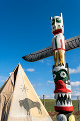 Colorful totem pole stands beside traditional teepee under bright blue sky