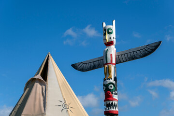 Colorful totem pole stands next to a traditional tent under a bright blue sky during a sunny day