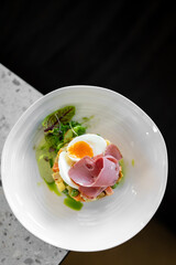 A gourmet dish featuring a soft-boiled egg, sliced ham, and a creamy vegetable salad with peas and carrots, garnished with fresh greens on a white plate