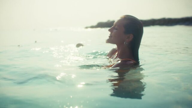 A serene woman gracefully floats and swims in the clear, calm tropical ocean water during a beautiful sunset. She embodies peace and relaxation on a luxurious summer vacation.