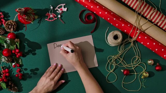 Top view of female hands writing a Christmas letter that begins Dear Santa, I want on a piece of paper surrounded by holiday decorations. Cozy Christmas mood and creative flat lay for holiday content.