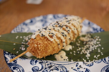 A deep fried food on a stick is topped with sauce and sesame seeds.