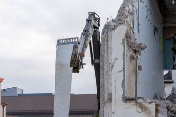 Excavator grab on a construction site, where a building is torn down, is holding a huge iron sheet against a cloudy sky. Wall of building besides, still lamps mounted on the concrete.