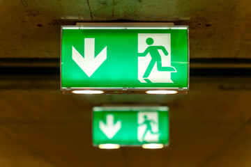 Two green emergency exit signs in a row, the background one blurry, below the ceiling of an underground garage., .