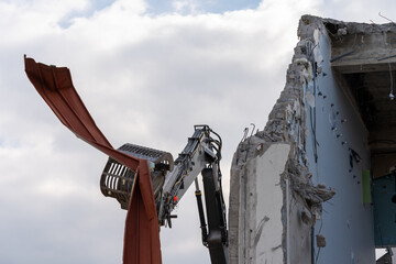 Excavator grab on a construction site, where a building is torn down, is holding a huge iron sheet against a cloudy sky, with a blue spot. Wall of building besides, still lamps mounted.
